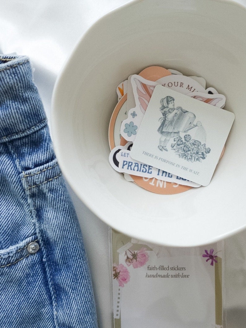 Puede incluir: Un taz&oacute;n blanco lleno de pegatinas coloridas con ilustraciones de flores, mariposas y una ni&ntilde;a regando plantas. Las pegatinas tienen mensajes inspiradores como "Let" y "There is purpose in the wait". El taz&oacute;n est&aacute; sobre una superficie blanca junto a una tarjeta que dice "pegatinas llenas de fe hechas a mano con amor".