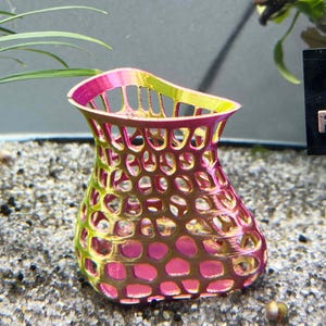 May include: A pink and yellow 3D printed vase with a mesh design. The vase is made of a lightweight plastic material and has a unique, organic shape.