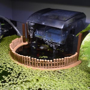 May include: An aquarium setup featuring a black filter system with water cascading into a small, round pond enclosed by a wooden fence. The pond is surrounded by green aquatic plants, with a large green leaf in the foreground.