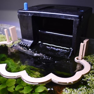 May include: A black aquarium filter with a blue component on top. Water flows from the filter into a decorative, light-colored, scalloped-edged structure. Green plants and moss surround the water feature.
