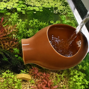 Peut inclure: Ornement d'aquarium en céramique marron avec de l'eau qui y coule. L'ornement a une forme unique avec une large ouverture et un bec. L'arrière-plan présente des plantes aquatiques vertes luxuriantes et un environnement d'aquarium clair.