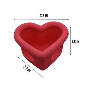 May include: A red heart-shaped container, 3.3 inches wide, 2.7 inches long, and 1.8 inches high. The container has a smooth, rounded rim and two small holes on the sides. The object is on a white background.