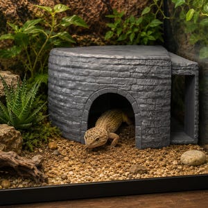 以下が含まれることがあります： レンガ模様のテクスチャとアーチ型の入り口を持つ、灰色の半円形爬虫類用シェルター。斑点模様のヒョウモントカゲモドキが、ガラスケース内の砂状の床材の上に置かれた構造物の入り口から、体の一部を外に出している様子。