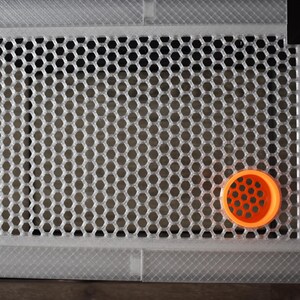 May include: A rectangular, silver vent cover with a honeycomb design. An orange, circular object with a similar pattern is on the lower right corner of the vent cover.