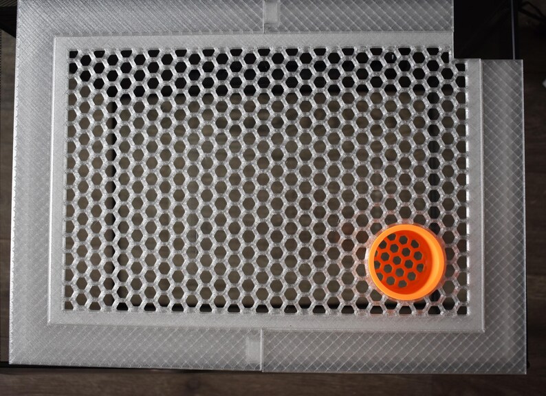 May include: A rectangular, silver vent cover with a honeycomb design. An orange, circular object with a similar pattern is on the lower right corner of the vent cover.
