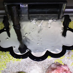 Puede incluir: Un deflector de filtro de acuario negro con forma de nube y una superficie plateada reflectante. El deflector est&aacute; colocado directamente debajo de la salida de agua de un filtro de mochila negro, capturando el agua que fluye y reduciendo la agitaci&oacute;n de la superficie.