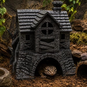 Peut inclure: Une cachette pour reptile gris foncé texturée en forme de maison à deux étages avec une cheminée, une fenêtre circulaire et des fenêtres condamnées. La structure présente une grande entrée voûtée à la base, où un gecko léopard tacheté se repose.