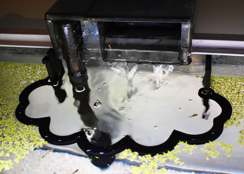 Puede incluir: Un deflector de filtro de acuario negro con forma de nube y borde festoneado, colocado directamente debajo de la salida de agua de un filtro de mochila negro. El deflector redirige el flujo de agua, creando una superficie lisa y reflectante.