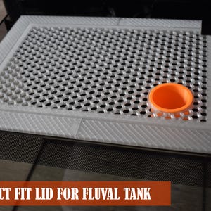 May include: A white, perforated lid for a Fluval tank with a diamond-patterned border. An orange, circular component sits on the lid. The text "PERFECT FIT LID FOR FLUVAL TANK" is displayed in a brown banner at the bottom.