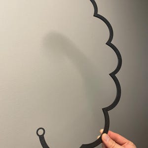 Puede incluir: Un marco negro con forma de nube, impreso en 3D, con una superficie texturizada. El objeto presenta una serie de bordes redondeados y festoneados en un lado y dos peque&ntilde;os puntos de fijaci&oacute;n circulares en los extremos superior e inferior.