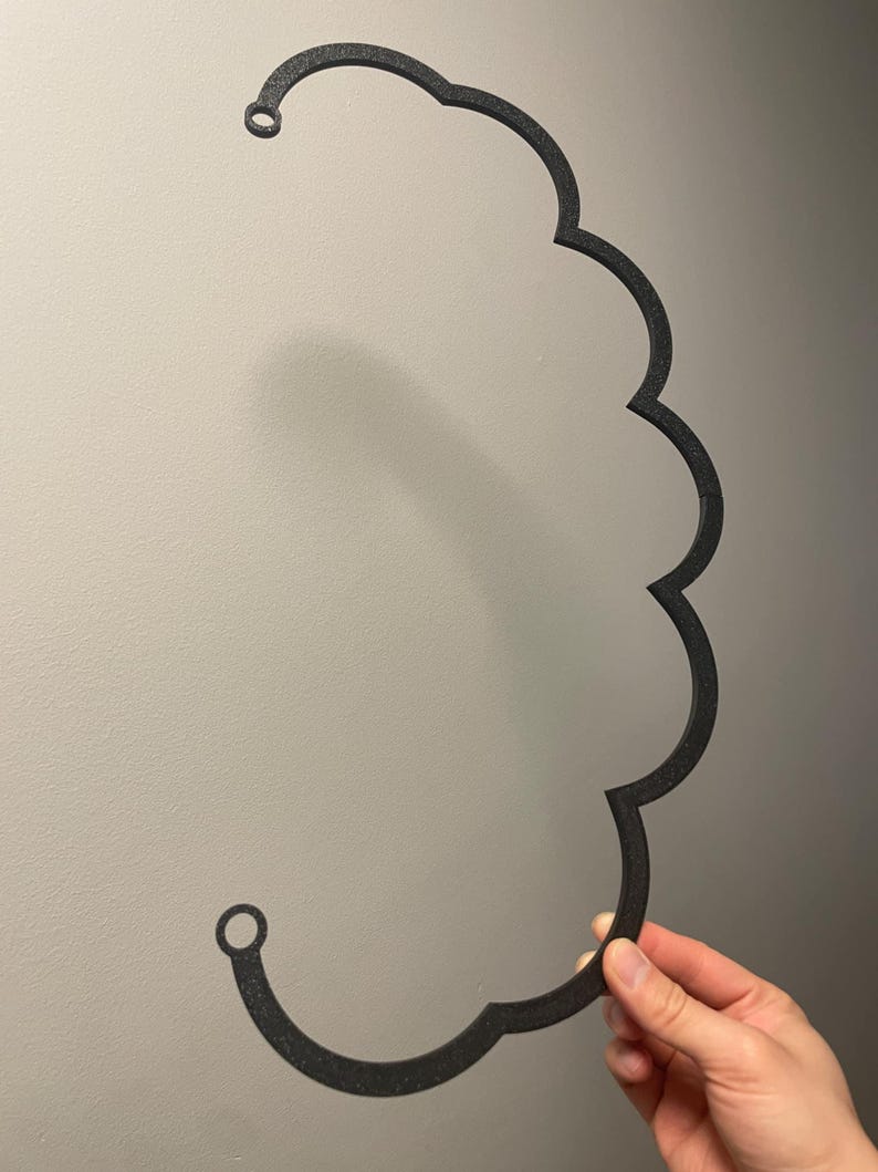 Puede incluir: Un marco negro con forma de nube, impreso en 3D, con una superficie texturizada. El objeto presenta una serie de bordes redondeados y festoneados en un lado y dos peque&ntilde;os puntos de fijaci&oacute;n circulares en los extremos superior e inferior.