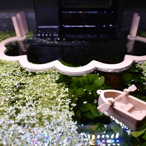 Puede incluir: Un corral flotante para plantas de acuario en forma de nube de color beige claro con borde festoneado y una peque&ntilde;a figura de bote de remos a juego con remos, ambos sobre la superficie del agua llena de plantas acu&aacute;ticas verdes y un filtro negro.