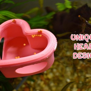 May include: A pink heart-shaped aquarium decoration with the text "UNIQUE HEART DESIGN". The decoration is in an aquarium with green plants and small orange objects inside. The design has two openings on the sides.