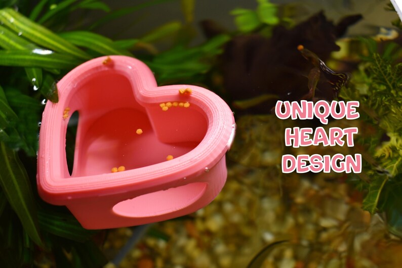 May include: A pink heart-shaped aquarium decoration with the text "UNIQUE HEART DESIGN". The decoration is in an aquarium with green plants and small orange objects inside. The design has two openings on the sides.
