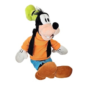 Może przedstawiać: Pluszowa zabawka Goofy'ego, postaci z kreskówek Disneya, jest pokazana w pozycji siedzącej. Goofy ma duży, beżowy nos, czarną głowę z zielonym kapeluszem i nosi pomarańczowy sweter, niebieskie spodnie i brązowe buty. Jego dłonie są białe.