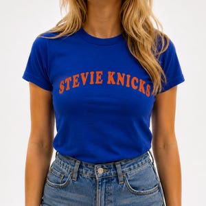 May include: A royal blue short-sleeved t-shirt featuring the text STEVIE NICKS printed in a bold, arched orange font across the chest. The shirt has a classic crew neckline and is paired with light blue denim jeans with a visible metal button.