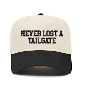 May include: Cream-colored baseball cap with a black brim. The cap features the embroidered text "NEVER LOST A TAILGATE" in bold, black letters. The cap has a curved brim and a button on top.