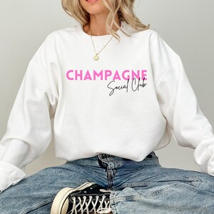 May include: White sweatshirt with the text "Champagne Social Club" in pink lettering.