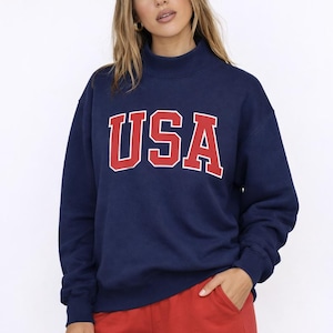 Könnte beinhalten: Marineblaues Sweatshirt mit dem Wort "USA" in roten Blockbuchstaben. Das Sweatshirt hat einen Rollkragen und lange Ärmel. Die Person trägt rote Shorts. Das Gesamtthema ist Freizeitkleidung.