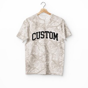 May include: A short-sleeved t-shirt in a khaki and white branch-like camouflage pattern. The shirt features the word CUSTOM printed in bold, black, arched capital letters across the chest. The garment is displayed on a wooden hanger against a white background.