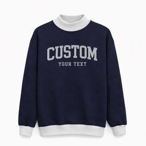 May include: Navy blue sweatshirt with a white turtleneck collar and cuffs. The front of the sweatshirt features the word "CUSTOM" in gray block letters above the words "YOUR TEXT". A comfortable and stylish garment.