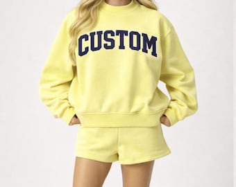 Op de afbeelding: Een felgele sweater met lange mouwen en ronde hals, gecombineerd met een bijpassende korte broek. Op de borst van de sweater staat het woord CUSTOM in grote, dikke, donkerblauwe blokletters. De set is gemaakt van een effen stof.