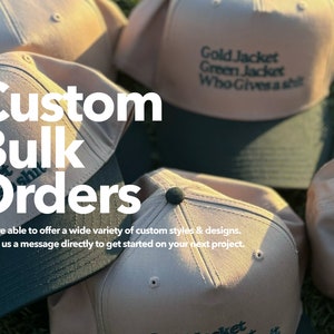 May include: A close-up of several beige baseball caps with green brims. The caps have different embroidered text, including "Gold Jacket", "Green Jacket", and "Who Gives a Shit". The text "Custom Bulk Orders" is superimposed on the image.