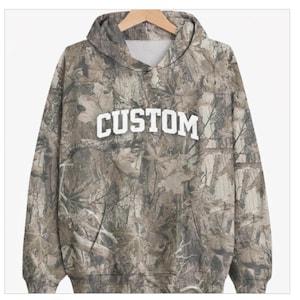 May include: A camouflage-patterned hoodie with the word "CUSTOM" in bold, white letters across the chest. The hoodie features a hood, a front pocket, and a drawstring. The fabric has a mix of brown, green, and tan tones.