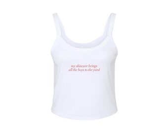 Op de afbeelding: Wit tanktop met een getailleerde zoom en de tekst "my skincare brings all the boys to the yard" in rood.