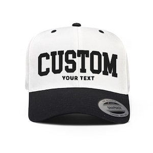 Pode incluir: Um boné snapback branco com aba preta e um botão preto no topo. A frente apresenta a palavra CUSTOM em letras pretas grandes, com as palavras YOUR TEXT impressas em letras pretas menores diretamente abaixo do texto principal.