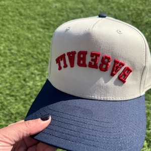 May include: A beige baseball cap with a navy blue brim. The word "BASEBALL" is embroidered in red on the front of the cap.
