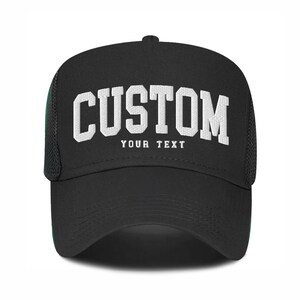 May include: Black trucker hat with white mesh back panel. The front of the hat has white text that reads "CUSTOM YOUR TEXT".