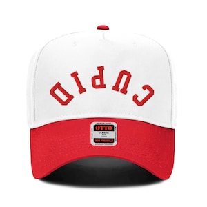 May include: White baseball cap with a red brim and the word "CUPID" written in red letters on the front.