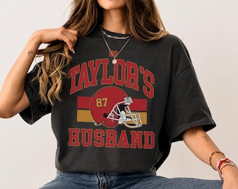 Könnte beinhalten: Ein anthrazitfarbenes Kurzarm-T-Shirt mit einem Grafikdruck, der den Text "TAYLOR'S HUSBAND" in roten Blockbuchstaben zeigt. Das Design enthält einen roten Football-Helm mit der gelben Nummer 87 vor horizontalen roten und goldenen Streifen.