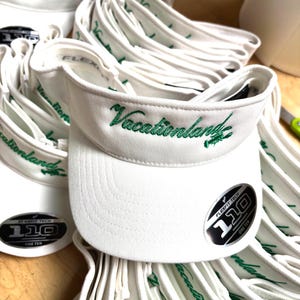 Pode incluir: Uma pilha de bonés brancos com bordado verde que diz "Vacationland" e uma etiqueta preta e prateada que diz "Flexfit Tech 110 One Ten".