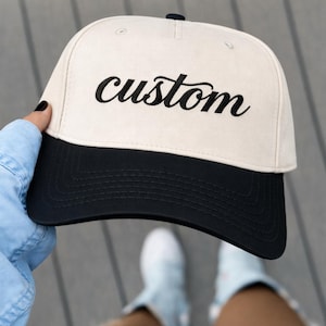 May include: A baseball cap with a cream-colored crown and a black brim. The word "custom" is embroidered in black script across the front. The hat is being held by a person wearing a light blue denim shirt.