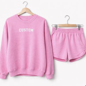 Puede incluir: Conjunto de sudadera y pantalones cortos rosas a juego. La sudadera tiene la palabra "CUSTOM" en letras blancas. Los pantalones cortos tienen cintura con cordón. Ambas prendas cuelgan de perchas de madera sobre un fondo blanco.
