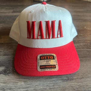 Pode incluir: Bon&eacute; de beisebol branco com aba vermelha e a palavra "MAMA" bordada em vermelho na frente. O bon&eacute; tem uma etiqueta lateral que diz "OTTO" e "Classic Fit OS FM" e "Mid Profile Maximum Comfort".