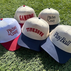 Op de afbeelding: Vijf baseballpetten met verschillende ontwerpen. De petten zijn wit met een gekleurde klep. De ontwerpen omvatten "Gameday", "Pitches Be Crazy", "Baseball" en "Play Ball".