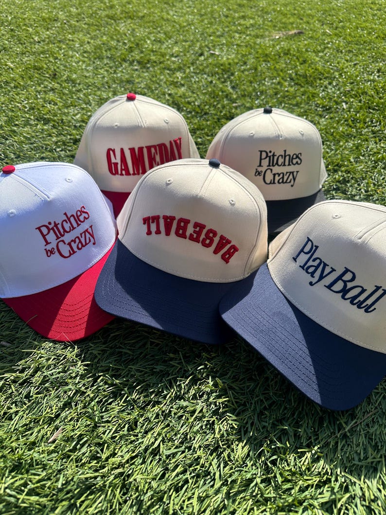 Op de afbeelding: Vijf baseballpetten met verschillende ontwerpen. De petten zijn wit met een gekleurde klep. De ontwerpen omvatten "Gameday", "Pitches Be Crazy", "Baseball" en "Play Ball".