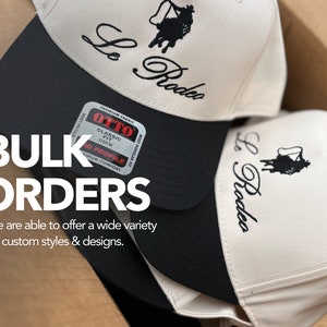 May include: Two white baseball caps with black brims and black embroidery that says "Le Rodeo". The caps are stacked on top of each other in a cardboard box. The text "BULK ORDERS" is in large white letters on the box. The text "We are able to offer a wide variety of custom styles & designs." is below the text "BULK ORDERS".