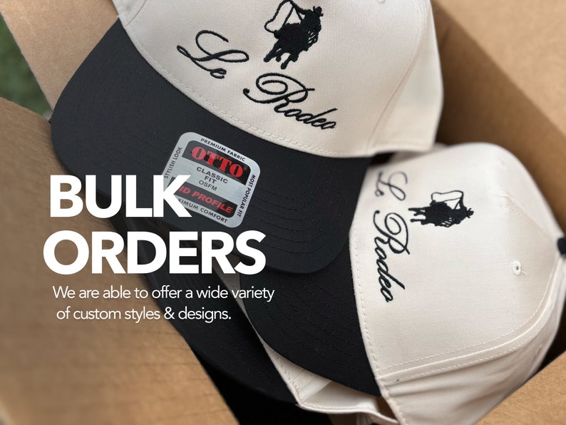 May include: Two white baseball caps with black brims and black embroidery that says "Le Rodeo". The caps are stacked on top of each other in a cardboard box. The text "BULK ORDERS" is in large white letters on the box. The text "We are able to offer a wide variety of custom styles & designs." is below the text "BULK ORDERS".