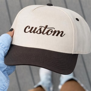 Könnte beinhalten: Beige-braune Baseballkappe mit dem Wort "custom" in dunkelbrauner Schrift auf der Vorderseite gestickt. Die Kappe hat einen gebogenen Schirm und einen Knopf oben.