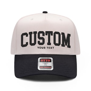 Pode incluir: Um boné de beisebol bicolor com copa bege claro e aba preta. A frente apresenta a palavra CUSTOM em letras pretas grandes acima do texto YOUR TEXT. Um adesivo retangular na aba diz OTTO CLASSIC FIT OSFM.