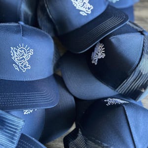 May include: A collection of navy blue trucker hats with white embroidered designs. The hats feature a mesh back and a curved brim. The embroidered design is a cherub with sun rays and lightning bolts.