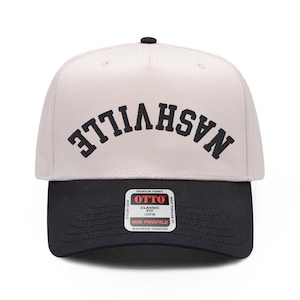 Pode incluir: Um boné de beisebol bege com aba preta e um botão preto no topo. A palavra NASHVILLE está impressa em letras maiúsculas pretas em uma orientação invertida e arqueada na frente. Um adesivo quadrado na aba diz OTTO.