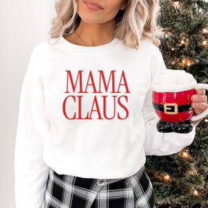 May include: White sweatshirt with the red text "MAMA CLAUS". A Santa-themed mug with a red coat design, black boots, and a white beard is held in the hand. The mug has a gold buckle. The background includes a Christmas tree.
