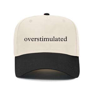 May include: A baseball cap with a cream-colored crown and a black brim. The word "overstimulated" is printed in black, centered on the front of the cap. The cap has a curved brim and a button on top.