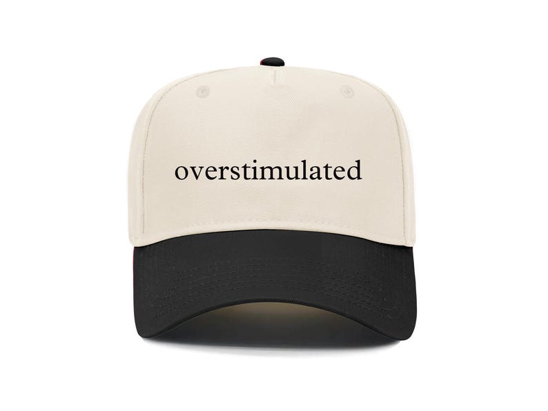 May include: A baseball cap with a cream-colored crown and a black brim. The word "overstimulated" is printed in black, centered on the front of the cap. The cap has a curved brim and a button on top.