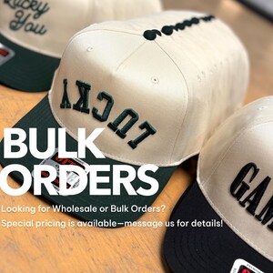 May include: Three baseball caps with cream-colored crowns and dark green or black brims. The caps feature embroidered text including 'LUCKY' in dark green and 'GAMEDAY' in black. Each cap has a structured front panel and a flat brim design.